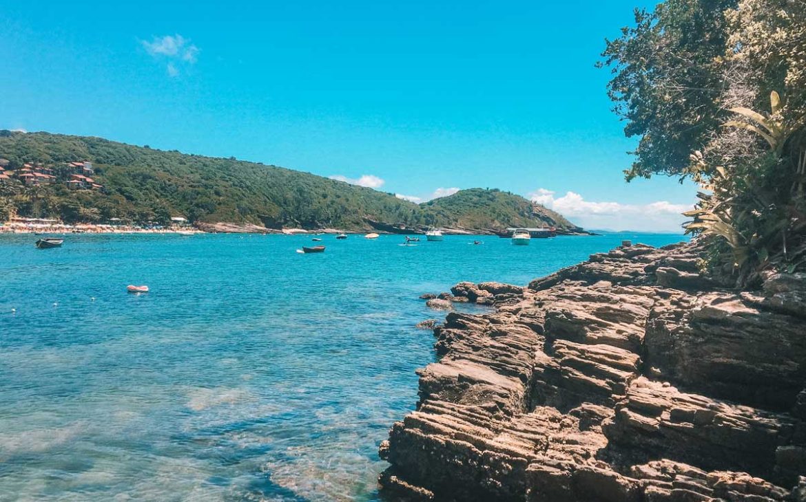 Formações rochosas à beira-mar em primeiro plano com águas cristalinas e tons de azul vibrante, cercadas por vegetação nativa e morros verdes ao fundo. Pequenos barcos flutuam ao longo da baía, transmitindo a atmosfera relaxante perfeita para quem procura onde ficar em Búzios e aproveitar paisagens naturais.