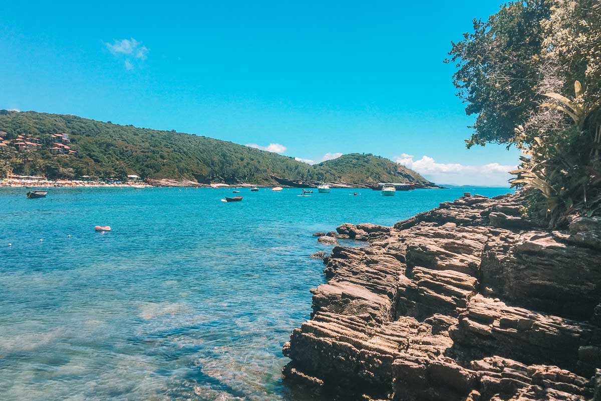 onde-ficar-em-buzios Formações rochosas à beira-mar em primeiro plano com águas cristalinas e tons de azul vibrante, cercadas por vegetação nativa e morros verdes ao fundo. Pequenos barcos flutuam ao longo da baía, transmitindo a atmosfera relaxante perfeita para quem procura onde ficar em Búzios e aproveitar paisagens naturais.